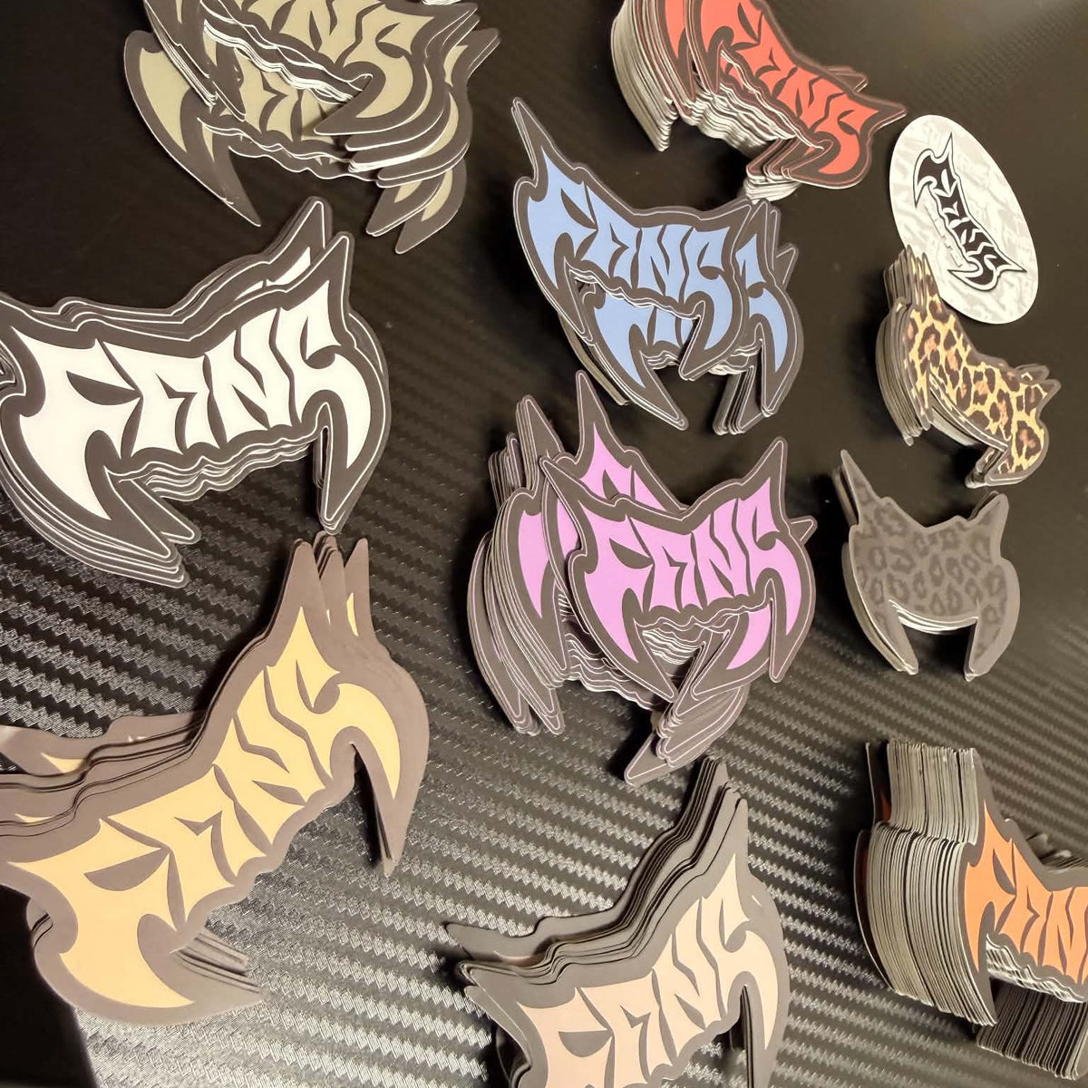 Multiple stacks of die-cut stickers featuring stylized animal head silhouettes with the word 'FANG' in bold gothic lettering, shown in assorted colors on a dark textured surface.