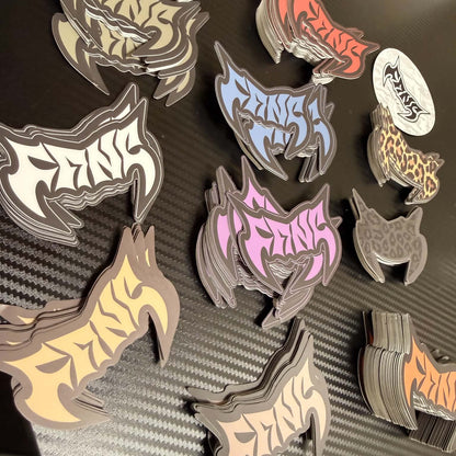 Multiple stacks of die-cut stickers featuring stylized animal head silhouettes with the word 'FANG' in bold gothic lettering, shown in assorted colors on a dark textured surface.